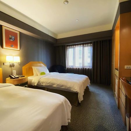 Ambassador Hotel Kaohsiung Buitenkant foto The photo shows a hotel room with two beds, each made up with white linens. The walls are painted in dark colors, while the bedding and furniture are lighter in tone. There is a nightstand between the beds with a lamp and telephone on it. A window wi