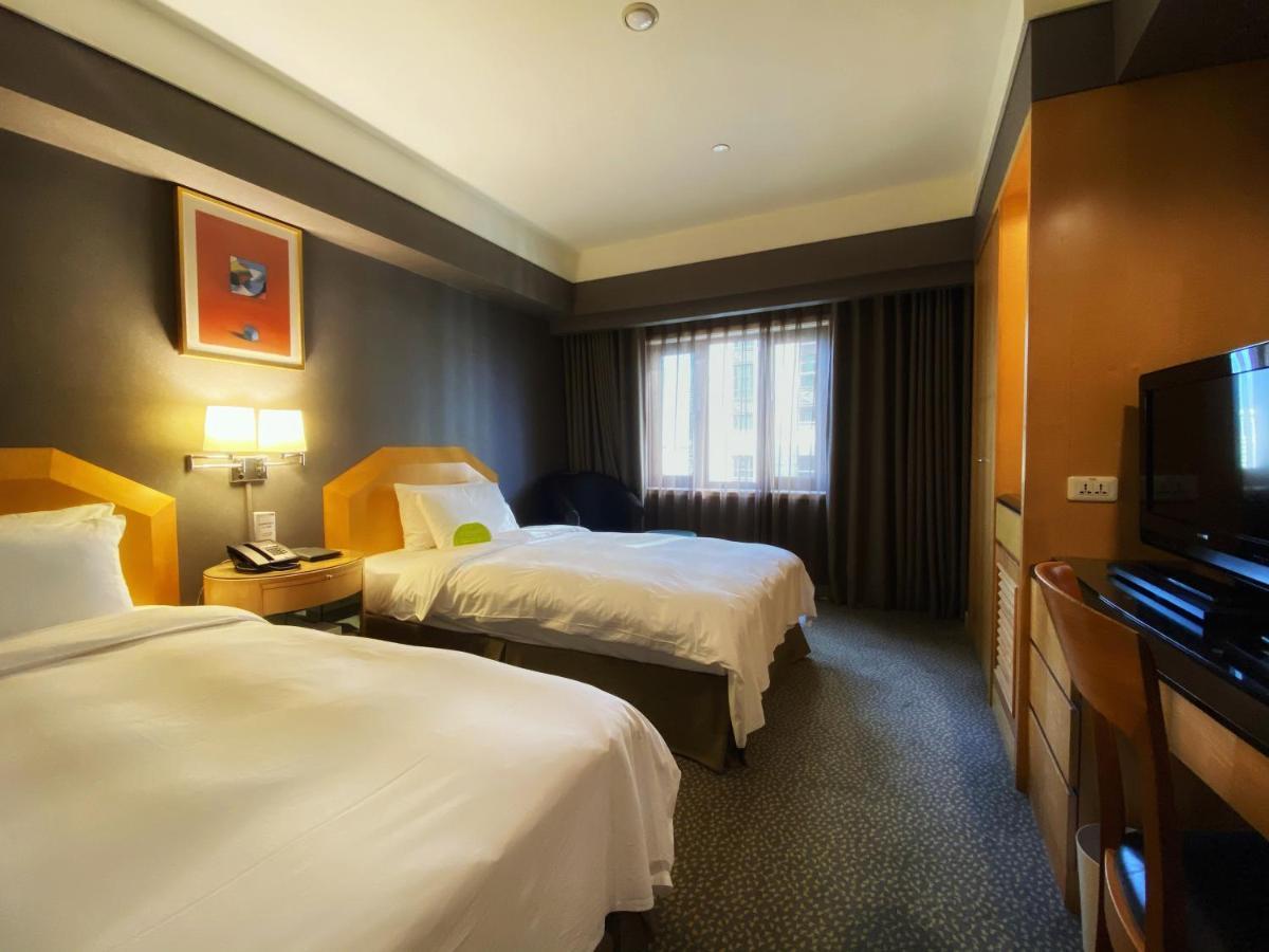 Ambassador Hotel Kaohsiung Buitenkant foto The photo shows a hotel room with two beds, each made up with white linens. The walls are painted in dark colors, while the bedding and furniture are lighter in tone. There is a nightstand between the beds with a lamp and telephone on it. A window wi