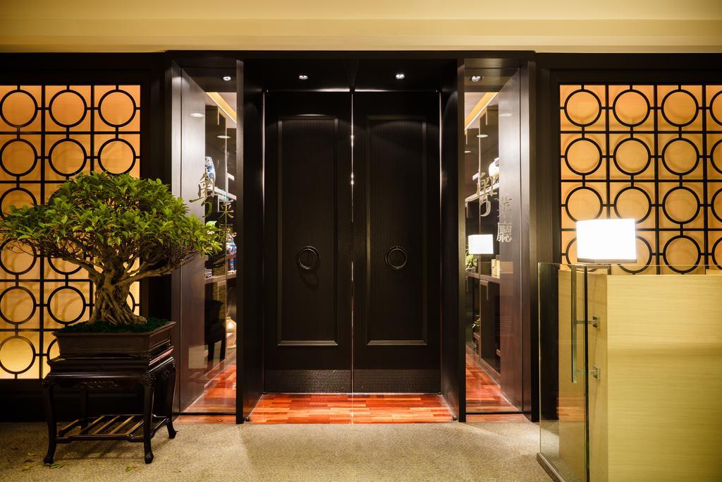 Ambassador Hotel Kaohsiung Buitenkant foto The photo shows an elegant entrance area featuring a set of large black double doors, which appear to be a main entryway. The walls surrounding the doors have a decorative design with circular patterns. On one side, there is a small tree in a planter