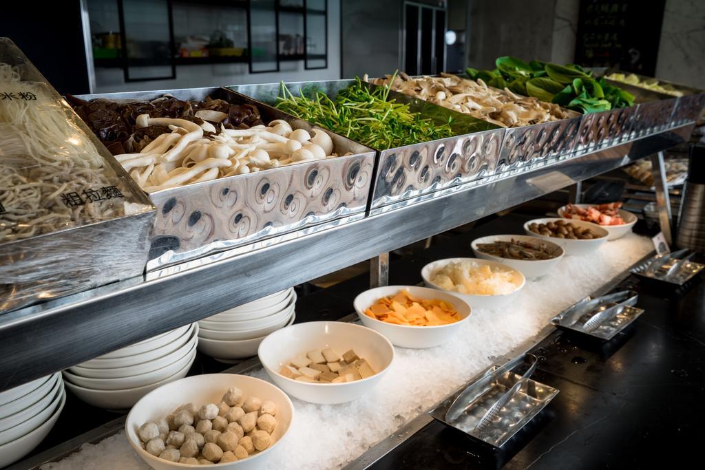 Ambassador Hotel Kaohsiung Buitenkant foto The photo shows a buffet or display of various food items, likely from an Asian cuisine setting. There are several metal trays filled with a variety of fresh vegetables, including greens and mushrooms. Below these trays, there are white bowls contain