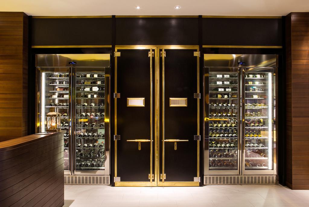 Ambassador Hotel Kaohsiung Buitenkant foto The photo shows a stylish, modern display area, likely in a restaurant or wine bar. It features large black double doors with gold trim at the center, flanked by glass refrigeration units on either side. The glass units are filled with various bottle