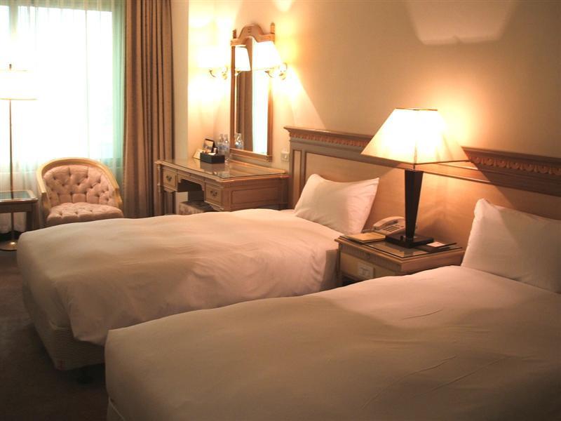 Ambassador Hotel Kaohsiung Buitenkant foto The photo shows a cozy hotel room with two single beds, each covered with white bedding. There is a lamp on a nightstand between the beds. A wooden dresser with a mirror is against the wall, and a comfortable armchair is placed nearby. Soft lighting 