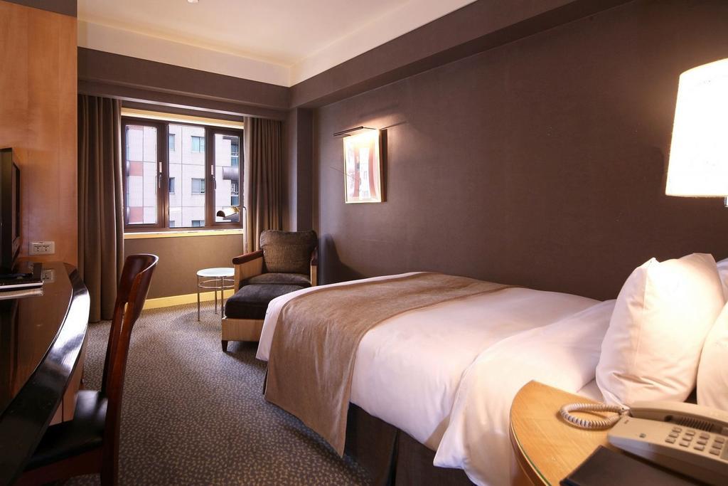 Ambassador Hotel Kaohsiung Buitenkant foto The photo shows a hotel room featuring a neatly made single bed with a light-colored blanket. There is a comfortable chair in the corner alongside a small round table. A large window allows natural light to enter the room, and the walls are painted i