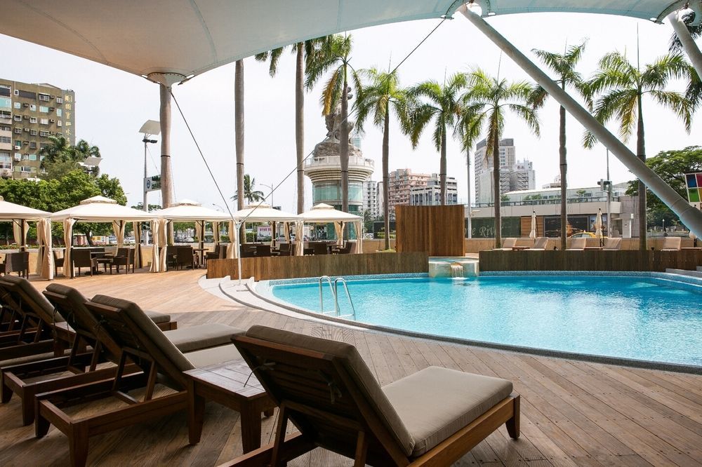 Ambassador Hotel Kaohsiung Buitenkant foto The photo shows a resort-style pool area. In the foreground, there are several wooden lounge chairs arranged around a sparkling blue pool. The pool has a gentle slope and is surrounded by wooden decking. Behind the pool, tall palm trees add to the tr