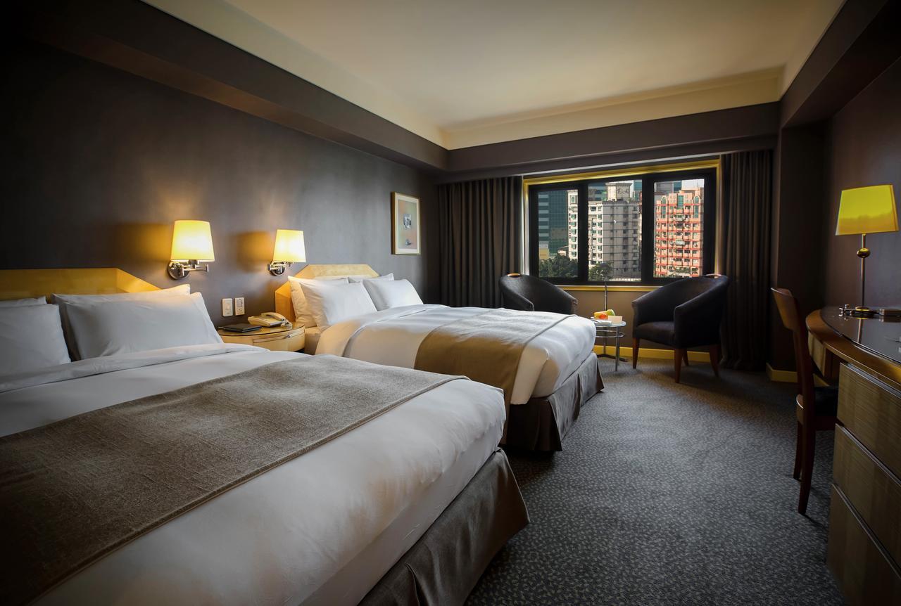 Ambassador Hotel Kaohsiung Buitenkant foto The photograph shows a hotel room featuring two double beds with white linens and light brown accents. The walls are painted a dark color, and there are bedside lamps beside each bed. A window offers a view of the outside, and there are two chairs po