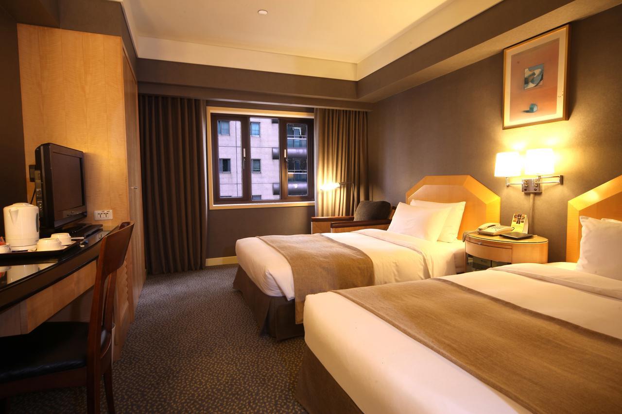 Ambassador Hotel Kaohsiung Buitenkant foto The photo shows a hotel room featuring two twin beds with neutral-colored bed linens. The room has a modern design, with warm lighting and wooden furnishings. There's a desk and chair positioned against one wall, along with a television. A large wind