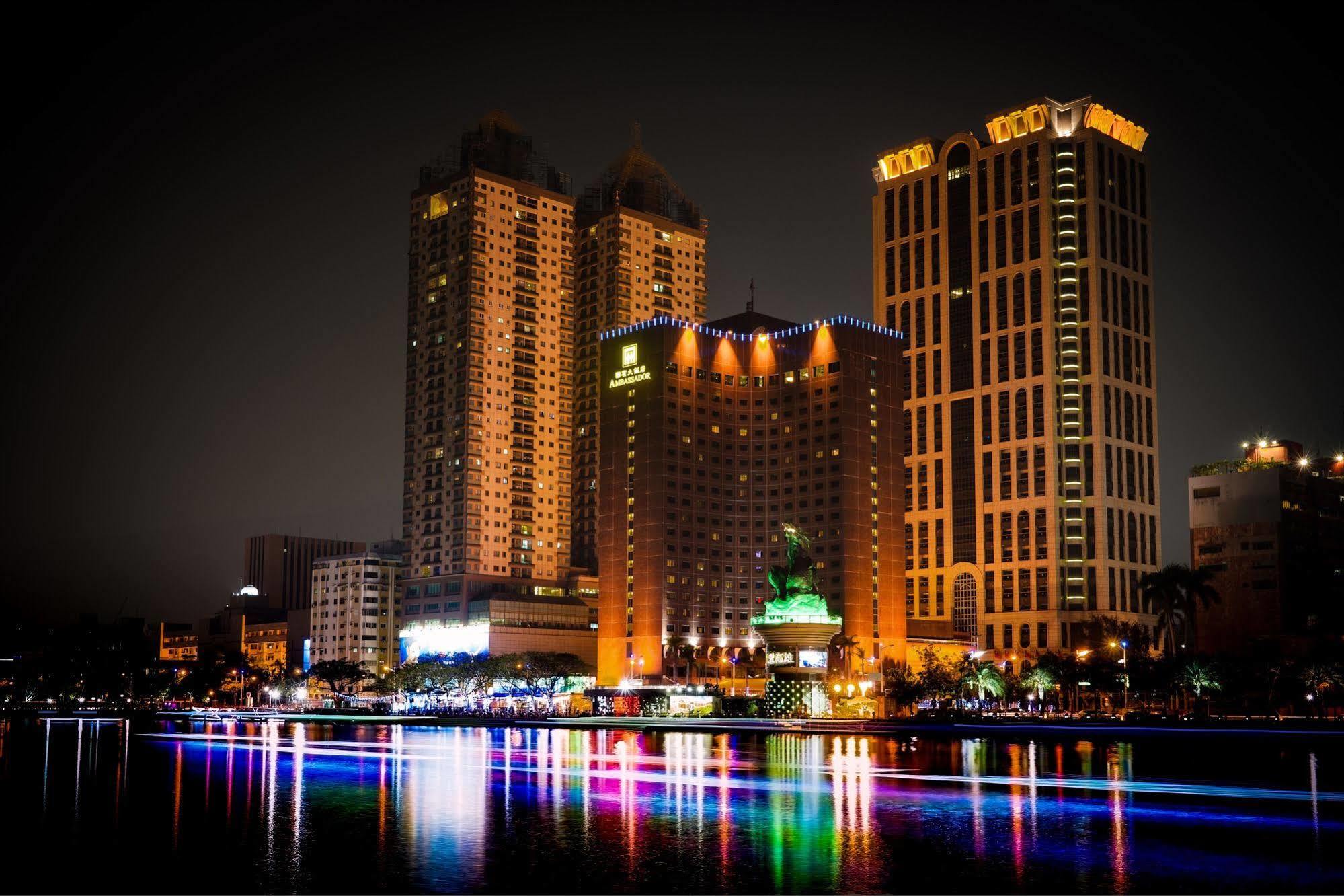 Ambassador Hotel Kaohsiung Buitenkant foto The photo depicts a vibrant nighttime cityscape featuring several tall buildings illuminated by colorful lights. Prominent structures include a large hotel and other high-rise buildings with illuminated facades. In front of these buildings, there app