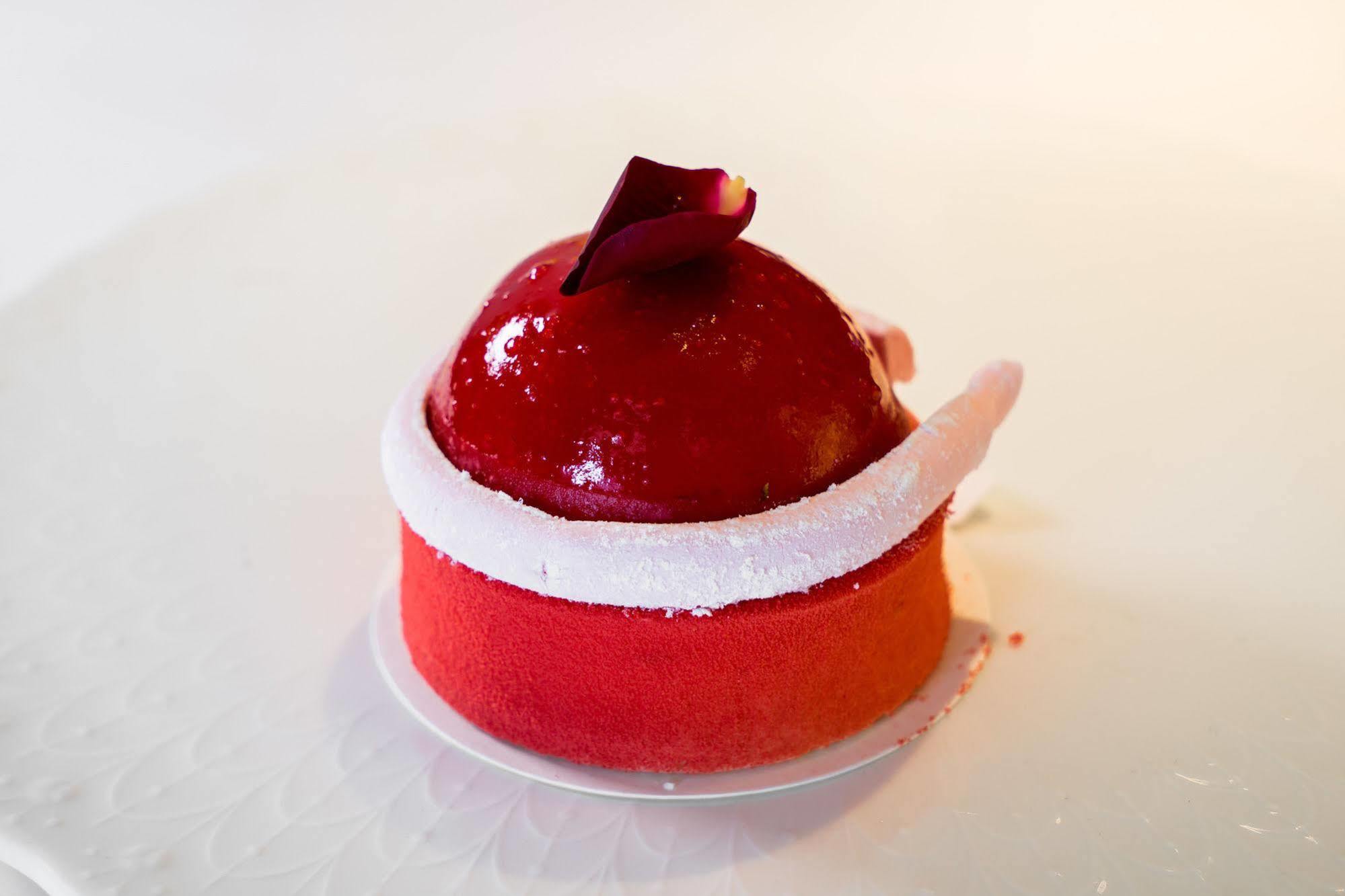 Ambassador Hotel Kaohsiung Buitenkant foto The photo shows a beautifully crafted dessert that appears to be a mousse or cake. It has a glossy red coating, shaped like a dome on top, with a small decorative rose petal placed on it. The base of the dessert features a gradient of colors, transit