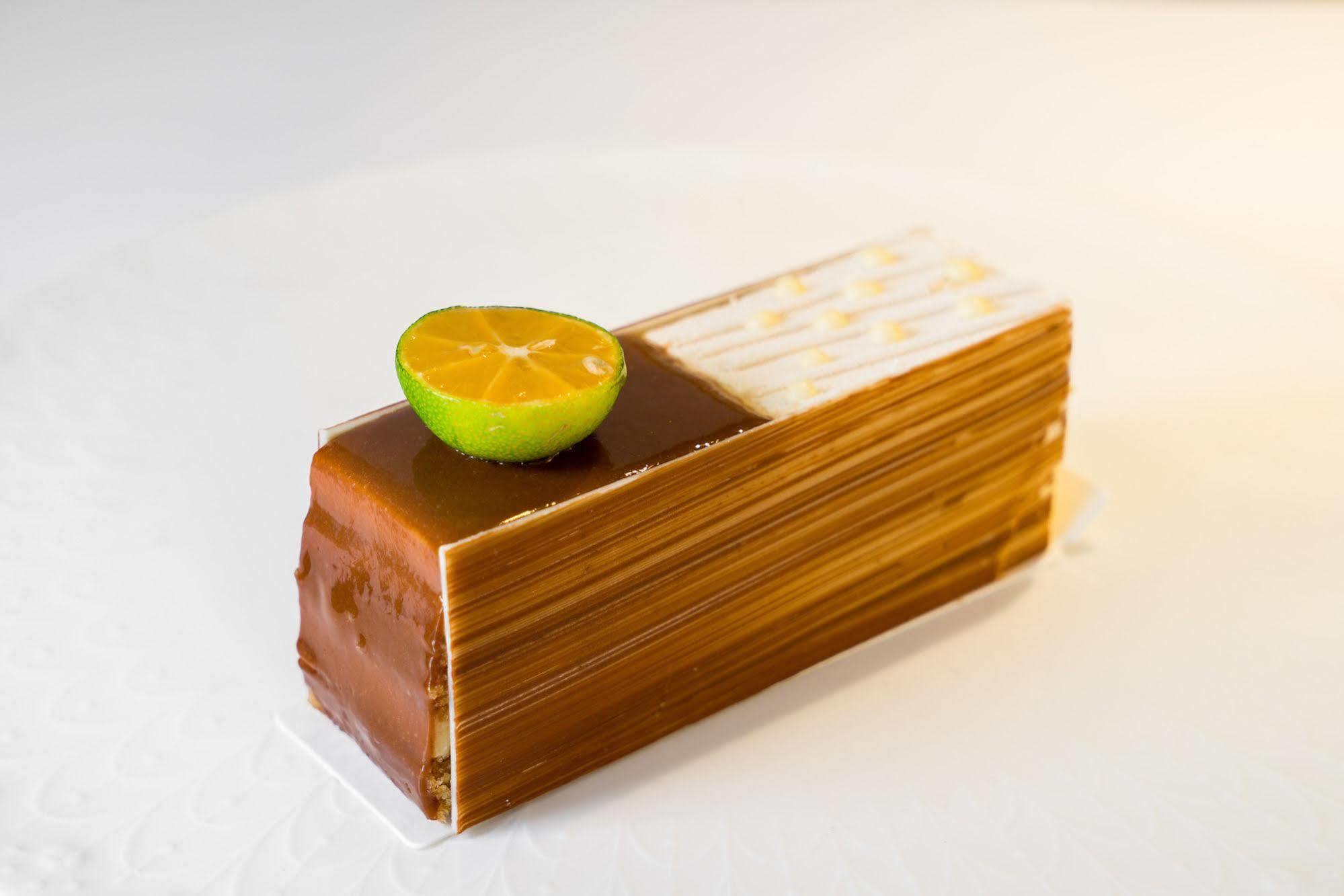Ambassador Hotel Kaohsiung Buitenkant foto The photo shows a dessert that is shaped like a rectangular block or loaf. It features a glossy, golden-brown exterior, likely made with layers of chocolate or caramel. Atop the dessert, there is a halved lime slice, adding a pop of color and suggest