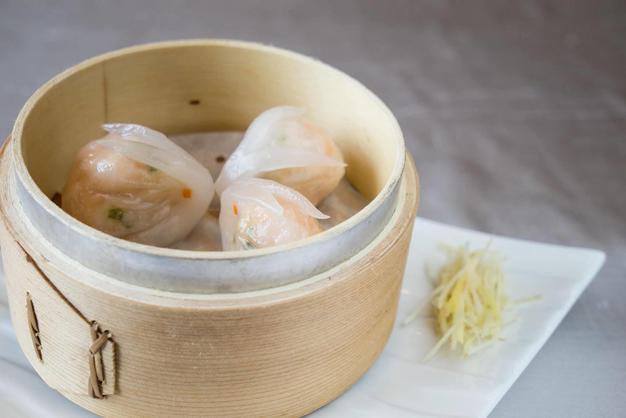 Ambassador Hotel Kaohsiung Buitenkant foto The photo shows a wooden steamer basket containing three pieces of dumplings, likely shrimp dumplings known as har gow. These dumplings are translucent, revealing bits of shrimp and vegetables inside. Next to the steamer, there is a small mound of sl