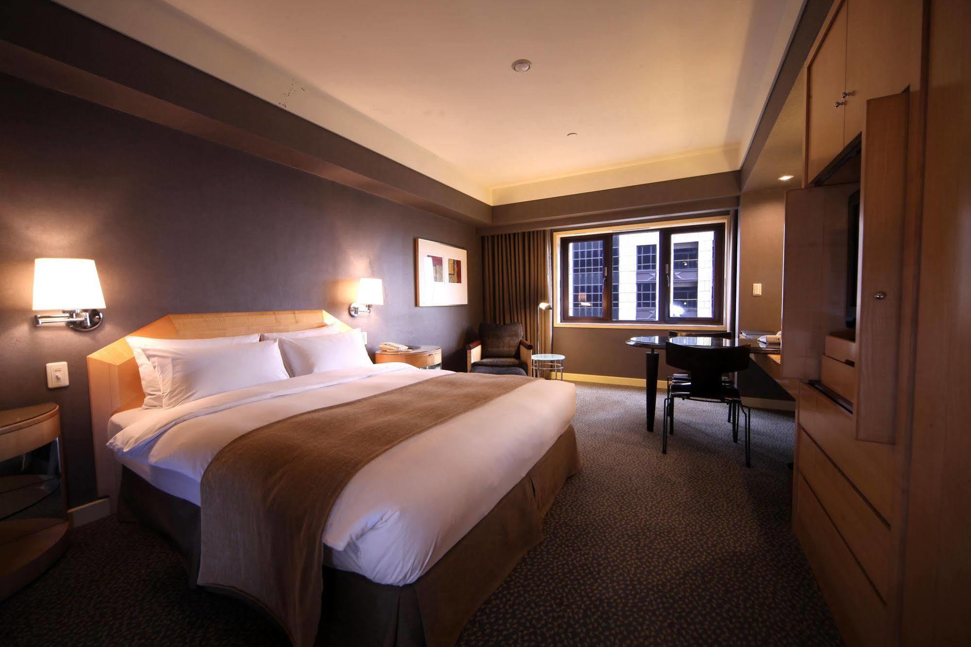 Ambassador Hotel Kaohsiung Buitenkant foto The photo shows a hotel room featuring a large bed with a white bedspread and a brown accent. There are nightstands on either side of the bed, each with a lamp. A chair is positioned near a window, which offers a view outside. A wooden desk and chair