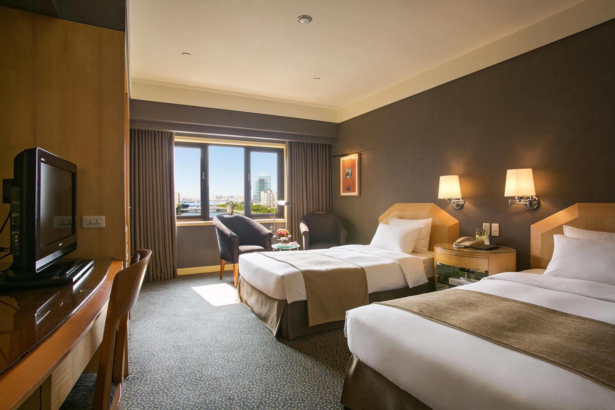 Ambassador Hotel Kaohsiung Buitenkant foto The photo shows a hotel room with two beds, one of which is a double bed and the other appears to be a single. The room has a modern design with a neutral color palette, featuring wooden furniture, a TV unit, and a small desk and chair. There are two