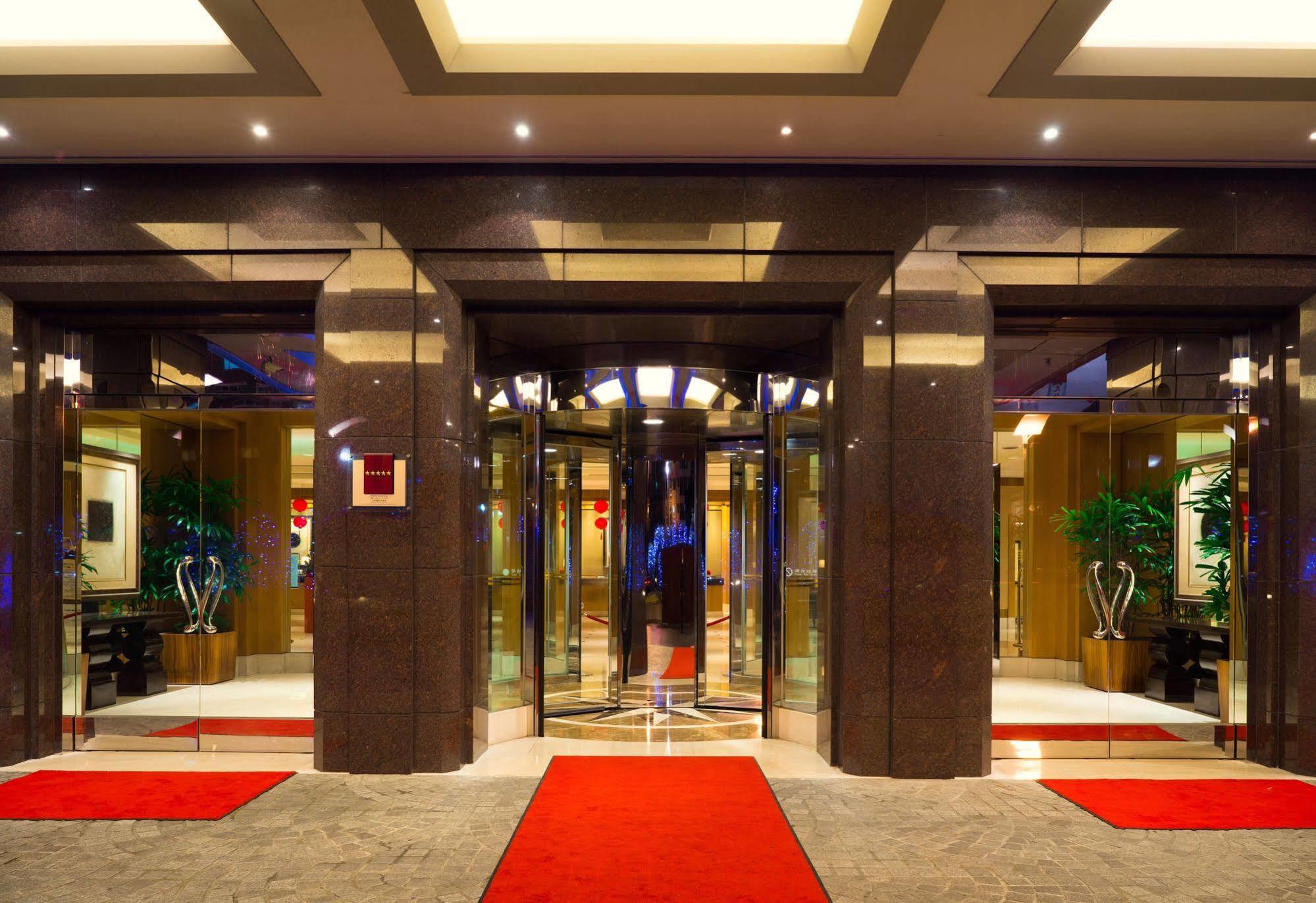 Ambassador Hotel Kaohsiung Buitenkant foto The photo shows the entrance of a modern hotel or building. It features large glass doors in a sleek frame, creating an inviting entrance. The floor is covered with a plush red carpet leading up to the doors. Inside, there are decorative plants on ei