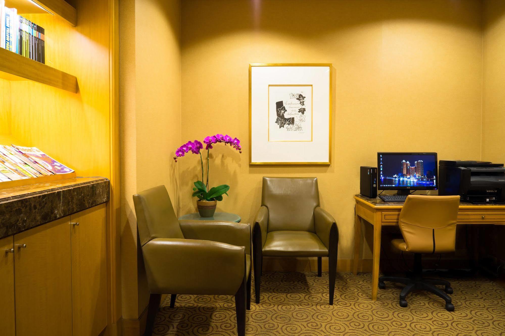 Ambassador Hotel Kaohsiung Buitenkant foto The photo shows a cozy corner of a room, which appears to be a waiting or lounge area. There are two stylish chairs positioned near a small table. A potted plant with purple flowers adds a touch of color and freshness to the space. On the wall, there