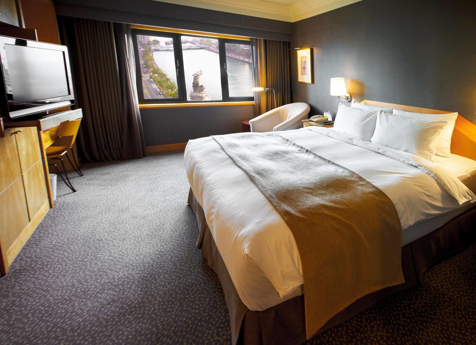 Ambassador Hotel Kaohsiung Buitenkant foto The photo depicts a modern hotel room featuring a spacious layout. In the center, there is a large bed with crisp white linens and a decorative brown throw blanket. To the side, there is a comfortable armchair and a small table. A television is mount