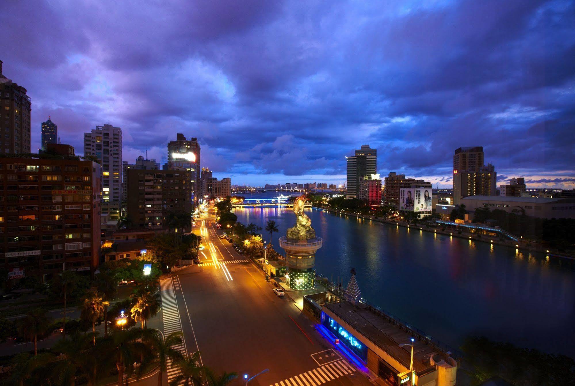 Ambassador Hotel Kaohsiung Buitenkant foto The photo depicts a cityscape during twilight. On the left, you can see a wide street lined with buildings, and the area is illuminated by streetlights. In the center, there is a body of water, potentially a river or canal, reflecting the lights from