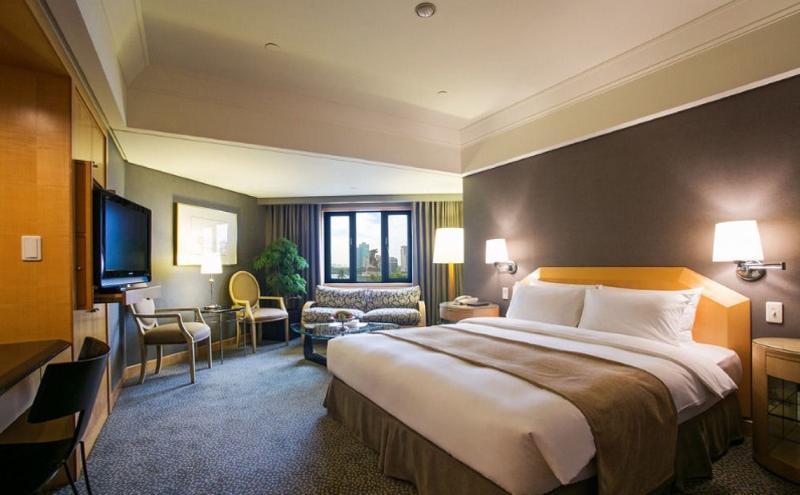 Ambassador Hotel Kaohsiung Buitenkant foto The photo shows a hotel room that features a large bed with white linens and a light brown throw blanket. There are two bedside lamps on either side of the bed. In the corner, there is a cozy seating area with a patterned sofa and two chairs. A telev