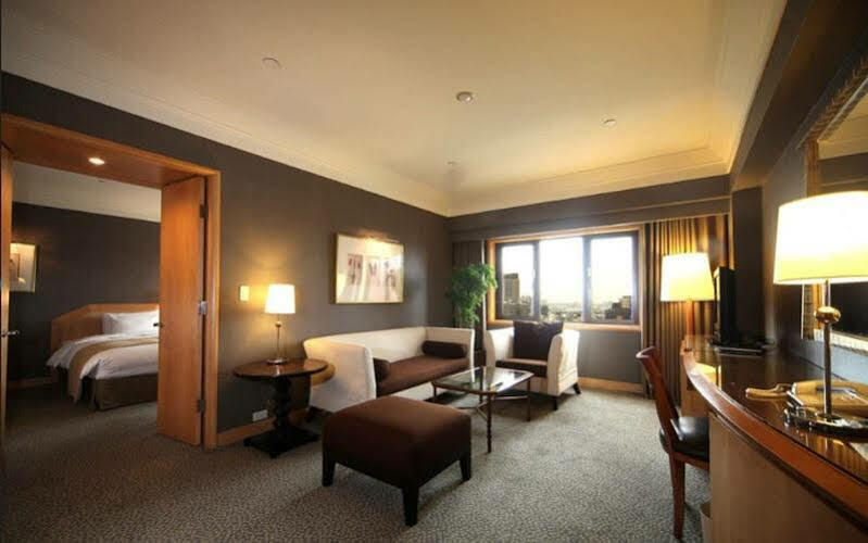 Ambassador Hotel Kaohsiung Buitenkant foto The photo shows a stylish hotel room with modern décor. There is a comfortable seating area featuring a white sofa and brown ottoman, along with a glass coffee table. A side table with a lamp is positioned next to the sofa. The room has a large windo